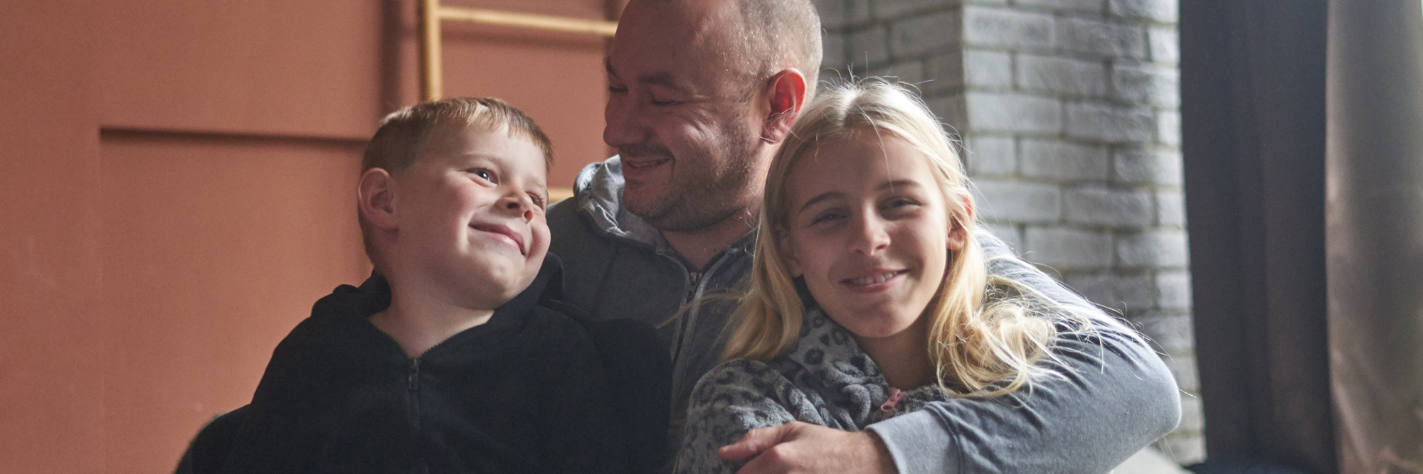 Man with short white hair hugging boy child with blonde hair and black shirt and girl with blonde hair, blue eyes wearing a flowered shirt.